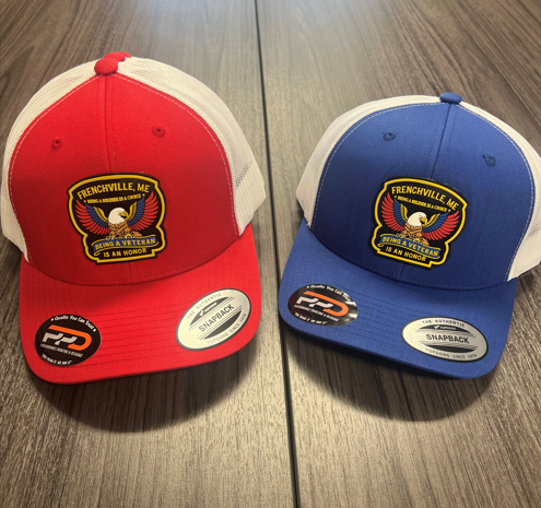 Two trucker-style caps on a wooden table: one red front with white mesh and a patch, and one blue front with white mesh and the same patch featuring an eagle.